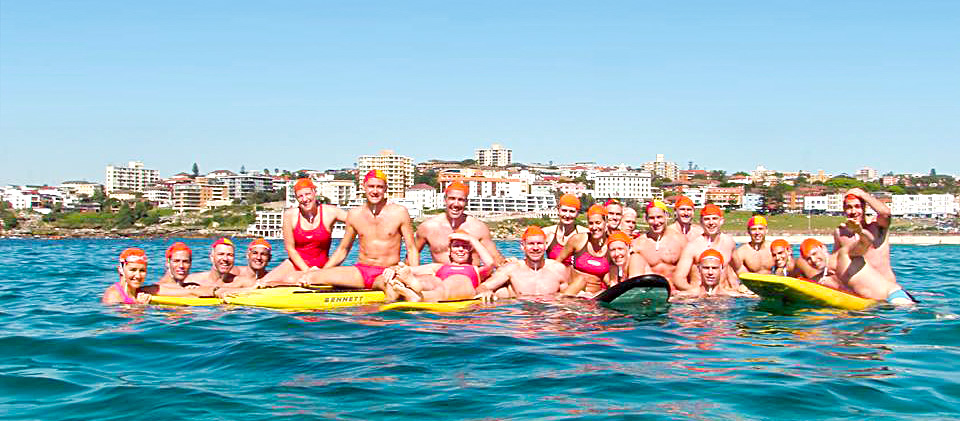 A group of around twenty men and women, all dressed in the red and yellow swimming uniforms of Australian Life Guards. They're pictured in the ocean, variously treading water, perched on rescue surfboards or holding onto the surfboards, all smiling at the viewer.